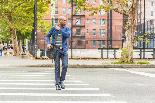 Young black man in New York crossing the street on the commute to work. Urban scene with playground and buildings on background. Lifestyle and millennials concepts