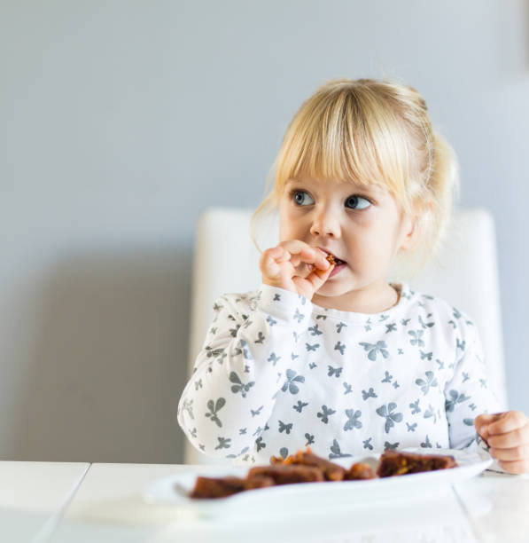 Little blonde toddler girl sitting at kitchen table and eating dinner stock photo