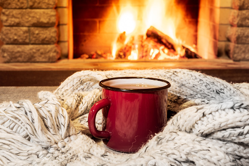 Red enameled mug for hot tea and cozy warm scarf near fireplace, in country house, winter vacation, horizontal.