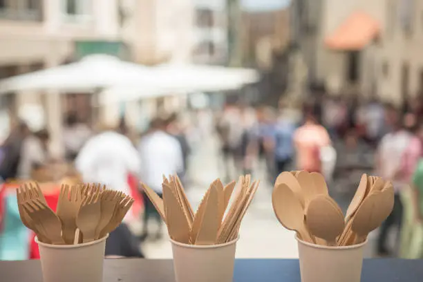 In the front of the photography is Disposable Wooden Cutlery – Eco Friendly Biodegradable Forks, Spoons, Knives. In the background is party on the street with plenty of people.