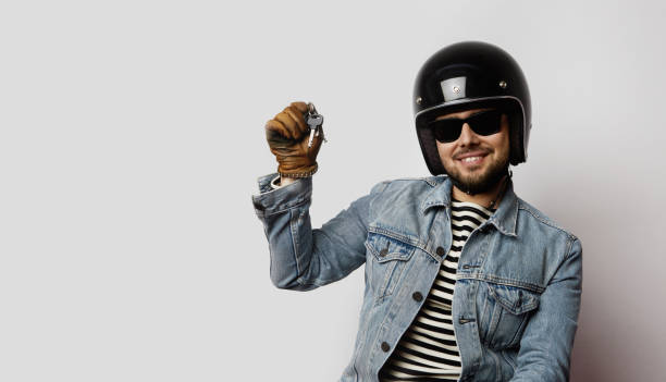 istock Young biker in a blue denim jacket pretending to ride a motorcycle isolated on white background.Man holding hand moto keys. 1061703370