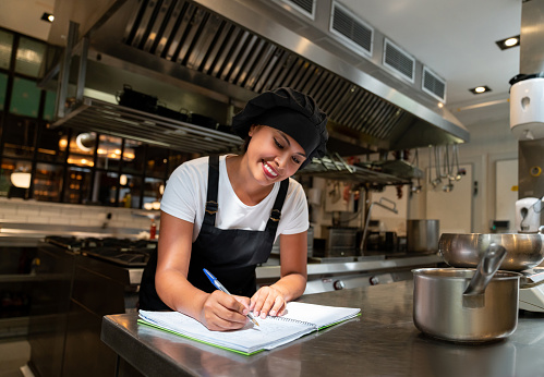 Portrait of a happy female chef working at a restaurant writing a recipe on a notebook and smiling - food and drink concepts
