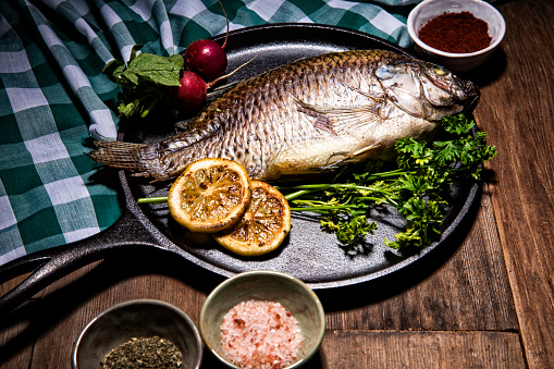 This is a photograph of gourmet Dorado fish on a metal skillet surrounded by colorful spices, lemons, parsley, and radishes in a full frame restaurant table setting. There are no people in the shot. The background is old retro wood with  a green checkered table cloth.