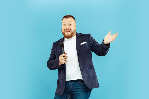 Young man with microphone on blue studio background, leading concept. Human emotions and facial expressions concepts.