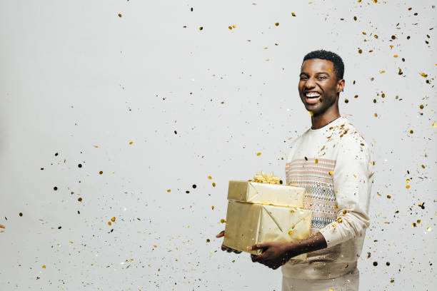 Portrait of a happy young man laughing and holding gifts as golden confetti are falling Portrait of a happy young man laughing and holding gifts as golden confetti are falling, isolated ion a gray studio background christmas excitement stock pictures, royalty-free photos & images