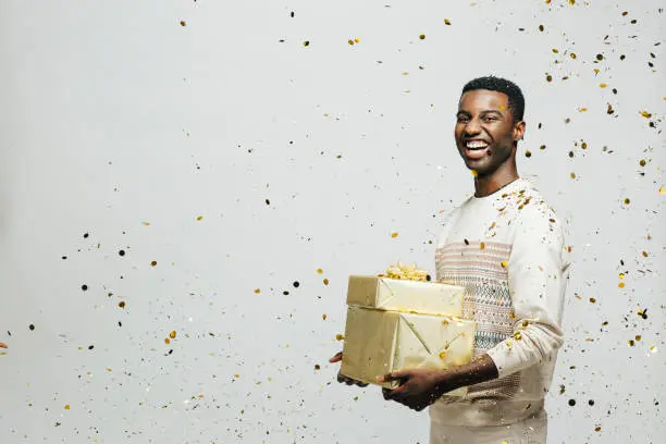 Photo of Portrait of a happy young man laughing and holding gifts as golden confetti are falling