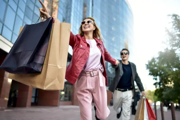 Photo of Front view of a casual couple of shoppers running in the street towards camera holding colorful shopping bags.