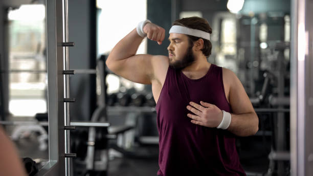 istock Funny fat man looking at mirror reflection gym and posing, pretending muscular 1061834258
