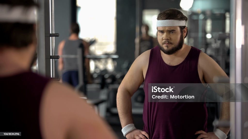 Unhappy overweight man looking at his mirror reflection in gym, diet and sport Gym Stock Photo