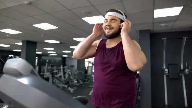 Photo of Fat man putting on headset and listening to music during jogging on treadmill