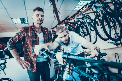 Consultant Shows Bicycle to Client in Sport Shop. Portrait of Young Shop Assistant Wearing White T-Shirt Helps in Mountain Bike Choosing. Happy Seller in Sport Store with Row of Bikes on Backround