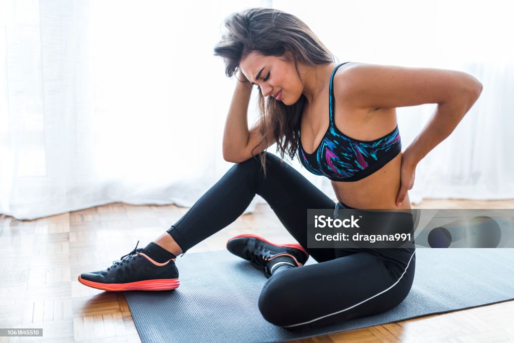 Woman suffering from lower back pain Woman suffering from lower back pain. Portrait of injured unhappy fit young beautiful woman sitting in sports club, touching her back after working out in class, suffering from backache, feeling pain, full length, side view Pain Stock Photo