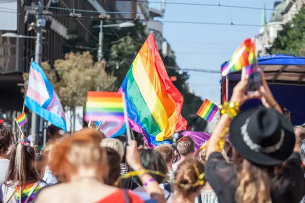 Photo of Crowd raising and holding rainbow gay flags during a Gay Pride. Trans flags can be seen as well in the background. The rainbow flag is one of the symbols of the LGBTQ community
