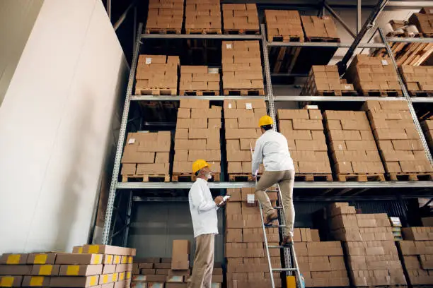 Photo of Senior worker using tablet and watching younger worker on ladders. Storage interior.