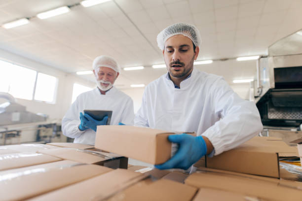 istock Man stacking boxes while other man standing in background and using tablet. Food factory interior. 1061870926