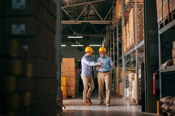 Workers walking in storage and pointing at boxes. In hands folder and tablet, on heads helmets.