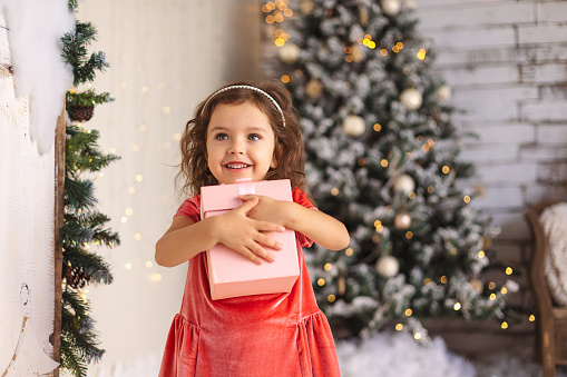 Cheerful little girl is hugging Christmas gift on Christmas tree and lights background.