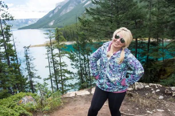 Blonde woman tourist wearing sunglasses and a jacket smiles and poses by Lake Minnewanka in Banff National Park in Alberta Canada