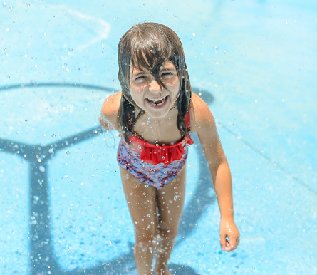 Little girl enjoys a splash pad or water park, she is getting wet and having fun in her bathing suit. She is adorable and full of joyful smiles and childhood exuberance. The best of summer. happy child with a fun childhood moment