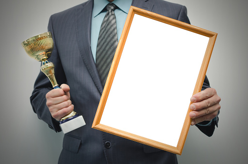 Man is holding a blank diploma or certificate frame with copy space and golden award trophy in the hands isolated on gray background.
