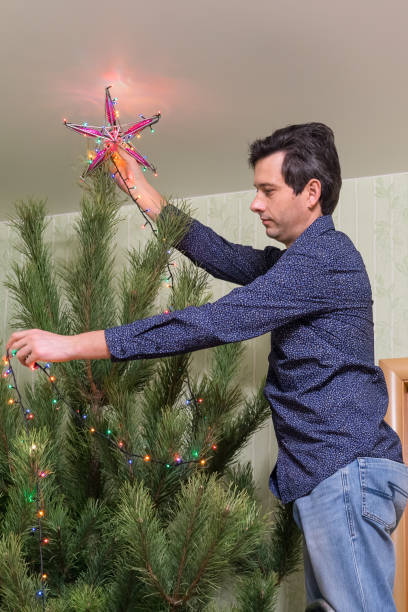 il bel uomo di mezza età decora un albero di natale con una stella - new year wall door decoration foto e immagini stock