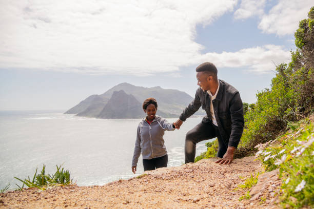 Young African couple walking on coastal path Man gives young woman a helping hand up slope on coastal walk chapmans peak drive stock pictures, royalty-free photos & images