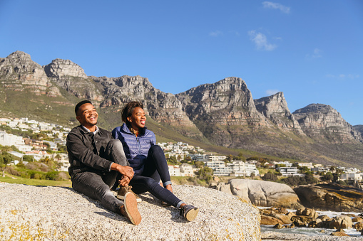 laughing young man and woman sit together in Camps Bay enjoying the view with Twelve Apostles behind