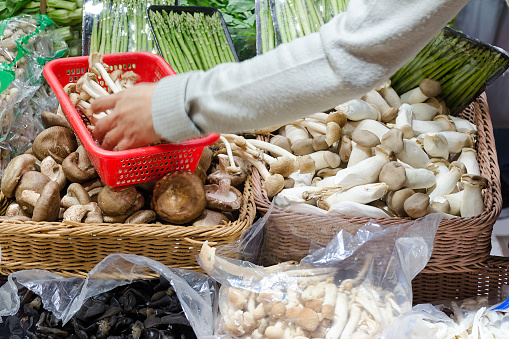 Stall with mushrooms and asparagus sprouts, hand fills the basket with mushrooms. Photo with blur in motion. Hong Kong, China.