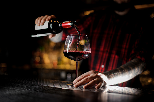 Professional bartender pouring a red wine from a bottle to a glass on the bar counter on the blurred background