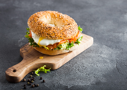 Fresh healthy bagel sandwich with salmon, ricotta and soft egg on vintage chopping board on stone kitchen table background