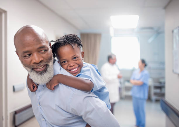 Father giving piggyback to son in hospital. Bearded father giving piggyback to son at children's hospital. Male nurse and ill boy are in corridor. They are enjoying at hospital. african father stock pictures, royalty-free photos & images