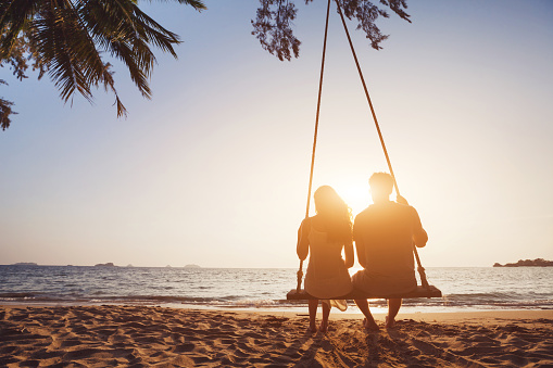 romantic couple in love sitting together on rope swing at sunset beach, silhouettes of young man and woman on holidays or honeymoon