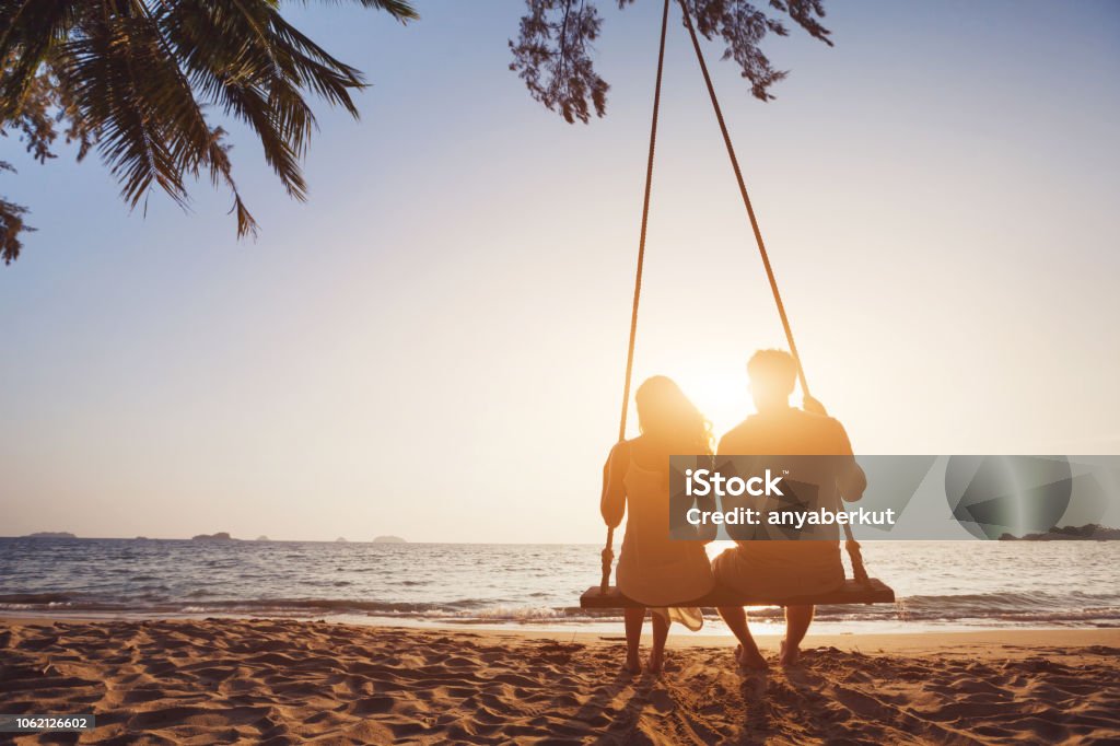 Honeymoon travel, silhouete of couple in love on the beach. romantic couple in love sitting together on rope swing at sunset beach, silhouettes of young man and woman on holidays or honeymoon Couple - Relationship Stock Photo