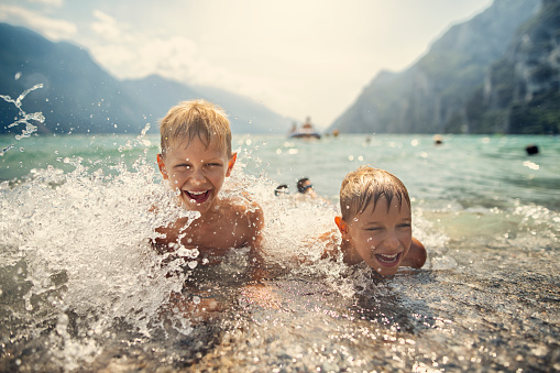 Brothers are having fun in lake in Riva del Garda, Italy. Kids aged 9 are lying on the beach and being splashed by lake waves. Boys are laughing and screaming.\nNikon D850