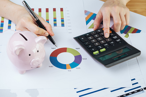 Hand holding pen reviewing pie chart and graph report with calculator and pink piggy bank on table, finance, money budget planning or investment asset allocation concept.