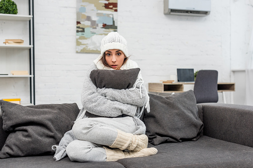 freezed mujer joven en ropa de abrigo sentado en sofá y abrazando el cojín en casa photo