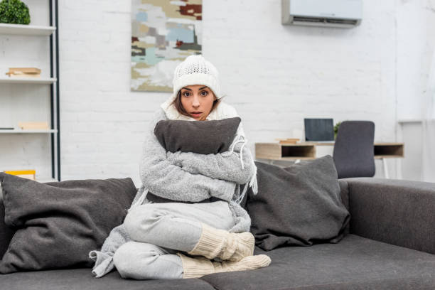 freezed mujer joven en ropa de abrigo sentado en sofá y abrazando el cojín en casa - frío fotografías e imágenes de stock