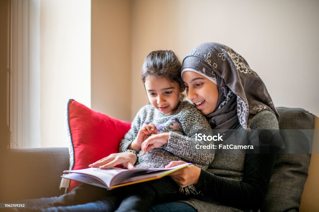 Big sister reads stories to her little sister A pre-teen girl wearing a hijab sits on a couch with her little sister on her lap and reads her a bedtime story. Her sister is engaged in the story. Family Stock Photo