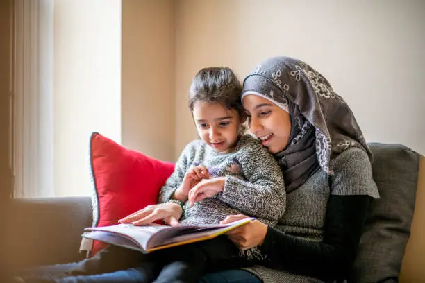 A pre-teen girl wearing a hijab sits on a couch with her little sister on her lap and reads her a bedtime story. Her sister is engaged in the story.