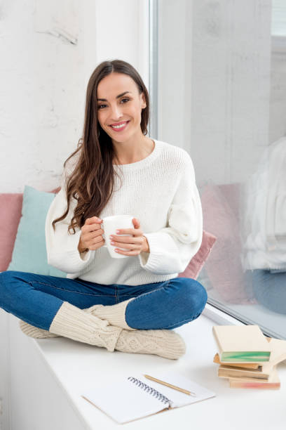 一杯のコーヒーの自宅の窓辺に座っているとカメラ目線で幸せな若い女 - window light window sill home interior ストックフォトと画像