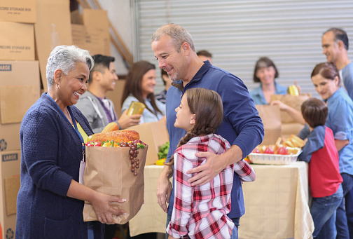 Multi-ethnic, mixed age group of volunteers work together at food bank at Thanksgiving.  They pack sacks and boxes of food for needy people in their community.  Senior woman gives sack full of groceries to single father and daughter.