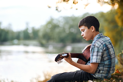the boy plays an acoustic guitar, sits on the Bank of the river, autumn forest at sunset, beautiful nature and the reflection of trees in the water