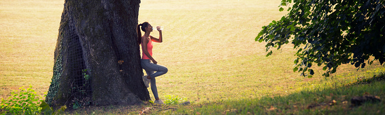 Young fitness woman drinking water after workout while leaning on ancient oak tree in summer.