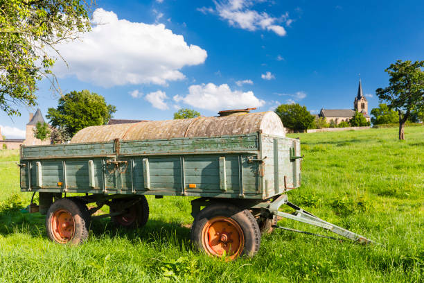 An old fertilizer trailer on a green meadow with a village in the background and blue sky.
