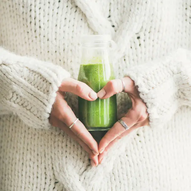 Winter seasonal smoothie drink detox. Female in sweater holding bottle of green smoothie or juice making heart shape with her hands, square crop. Clean eating, weight loss, healthy dieting food concept