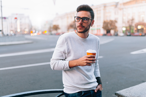 Hipster man standing with takeaway coffee, smiling plesantly, walking on th city street. Happy carefree handsome guy in eyewear. Millennial generation