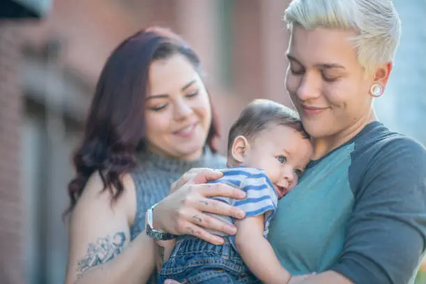 A smiling blonde lesbian looks down at her cute baby while holding him. Her wife looks at their son while placing a hand on his back.