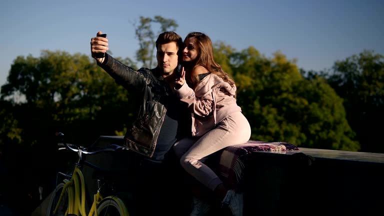 Beautiful young couple taking selfie by mobile phone on street while girl sitting on the parapet. Beautiful couple selfie, love and fun concept. Posing, the girlshowing peace sign. Trees around