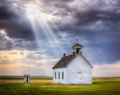 An old abandoned church with sun rays beaming down from the clouds. There is also a small outhouse beside the church. Both are in an open field with nothing around.