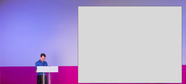 istock Presenter at Podium with Blank Presentation Screen. Copy Space for Text. Lecture on Stage at Business Education Conference Hall. Technology Training Seminar Event with Audience. 1062382528